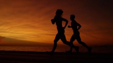Genç kadın ve adam deniz kenarında nefes kesen bir akşam bir akşam jog için gidin. Çarpıcı turuncu günbatımı önünde deniz boyunca çift jogging awesome yavaş hareket çekim