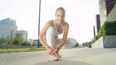 Slow Motion, Lens Flare, Portrait: Beyaz fitness giysileri mutlu kadın ayakkabılarını bağlar ve güneşli bir sabah şehir ortamında kamera geçmiş çalışır. Fit kız onu maraton eğitimi için hazırlar.
