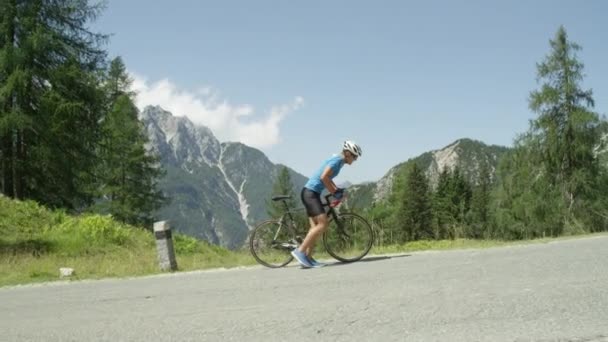 激しい山岳レース中に彼のクールな道路の自転車のペダルを踏んで運動若い男性 日当たりの良い山のプロロードサイクリストトレーニング 夏の自然の中で絵のように美しい自転車に乗る スローモーション映像 — ストック動画