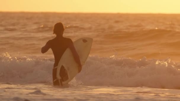 スローモーション 黒ウェットスーツで認識できないサーファーは 壮大な夕日に大きなクラッシュ波をサーフィンに行きます 夕暮れ前にガラス張りの海の波をサーフィンするために深海の水の中を歩く気楽なサーファーの男 — ストック動画