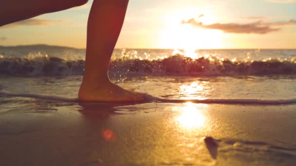 スローモーション クローズアップ アクティブな女性は 黄金の日の出に海岸に海の波が転がり込むように暖かい砂の上にジョギング 息をのむような夕日が砂の中でジョギングする認識できない女性を照らす — ストック動画