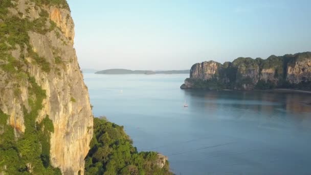 在泰国阳光明媚的旅游海滩附近 沿着巨大的石灰岩悬崖飞行 覆盖着郁郁葱葱的热带植物 巨大的岩溶地层在接近遥远海滩的蓝色海浪中翱翔 — 图库视频影像