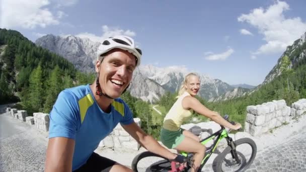 スローモーションSelife笑顔のカップルは 美しい夏の日に山道を自転車に乗って笑顔 山でサイクリングを楽しむ魅力的な新婚夫婦 カメラに手を振り Eバイクをペダリングする少女 — ストック動画