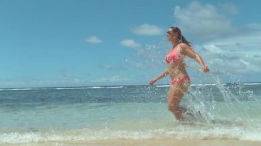 SLOW MOTION CLOSE UP: Happy young woman in pink swimsuit running on sandy beach, splashing ocean water. Smiling girl enjoying fun vacation in tropical island on sunny day. Exotic travel destination