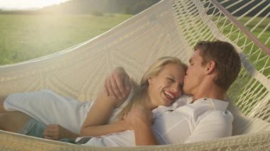 Slow Motion, Lens Flare, Portre: Neşeli adam romantik askıda yatarken sarışın kadını alnından nazikçe öpüyor. Doğada güneşli bir günde hamak içinde snuggling Güzel erkek arkadaşı ve kız arkadaşı.