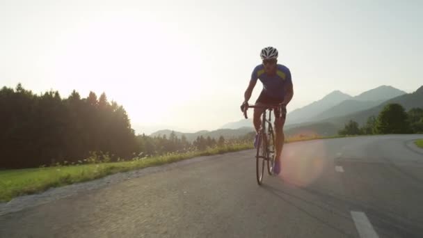 激しい山岳レース中に彼のクールな道路の自転車のペダルを踏んで運動若い男性 日当たりの良い山のプロロードサイクリストトレーニング 夏の自然の中で絵のように美しい自転車に乗る スローモーション映像 — ストック動画