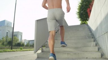 Yavaş Hareket, Düşük Açı, Lens Flare: Tanınmayan gömleksiz adam şehirde merdiven bir uçuş kadar jogs. Gri şort lu ve mavi spor ayakkabılı beyaz adam şehirde güneşli bir sabahta çalışıyor..