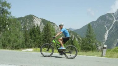 Slow Motion Close Up: Neşeli genç bisikletçi güneşli yaz gününde kolayca bir dağ yolunda elektrikli bisiklet sürme. Adam onun e-bisiklet üzerinde kolaylıkla bisiklet. Fit erkek binici zahmetsizce dik yol yükselen