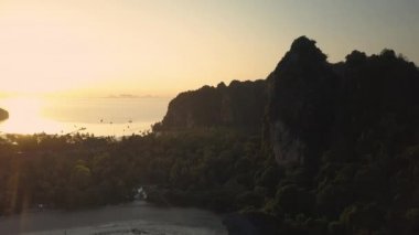 AERIAL, LENS FLARE: Beautiful golden sunbeams shine on tropical sandy beach and large cliff on remote island. Flying over tranquil exotic beaches and dense vegetation on tropical island at sunset.