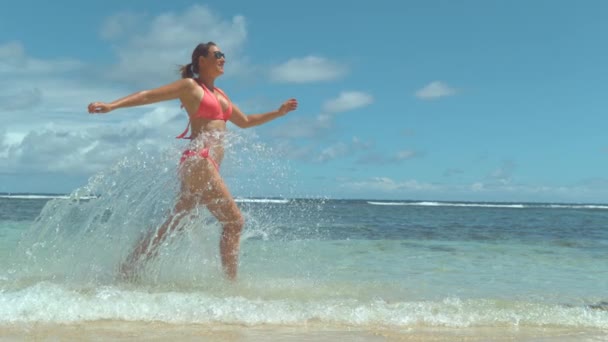 スローモーションクローズアップ 海水をはねかねて 砂浜で走るピンクの水着を着た陽気な若い女性 晴れた日に熱帯の島で楽しい休暇を楽しんで笑顔の女の子 エキゾチックな旅行先 — ストック動画