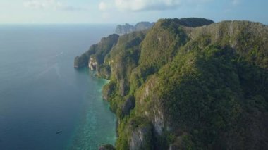 AERIAL: Flying away from enormous limestone islands surrounded by tranquil ocean water. Awesome shot of tropical green island cliffs and small tourist boats sailing near the picturesque landscape.