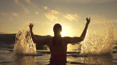 Slow Motion, Close Up, Lens Flare: Eğlenceli kız okyanus kıyısındaki sonsuzluk havuzundan ellerini kaldırırken, parlak su damlacıkları havada uçar. Yaz akşamı güneş ışınları su sıçrayan genç kadın üzerinde parlıyor