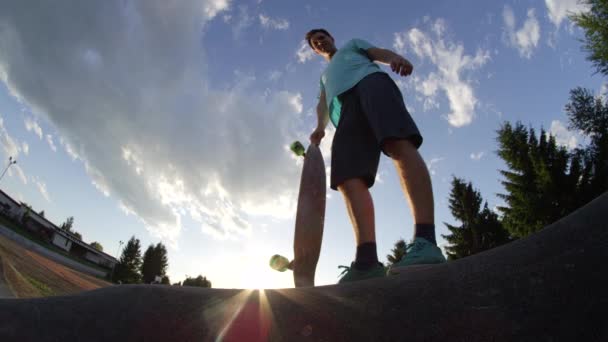 スローモーションローアングルレンズフレア 彼の実行を開始するために暖かい夏の風の中で待っているクールなスケートボーダー 彼のロングボードを保持し スケートパークで暖かい夕日の光の中に立っている若い男性をフィット — ストック動画