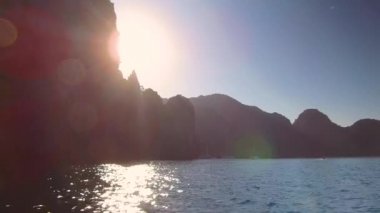 Pov, Lens Flare: Devasa kayalıkların karanlık ana hatları, parlak yaz güneş ışınlarıyla aydınlatılmış mavi okyanusa bakmaktadır. Kireçtaşı karstları geçmiş yelken ve sakin tropikal koyda demirlemiş turistik gemilere doğru.