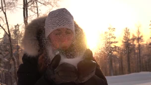 慢动作特写 年轻女子在冬天的夕阳下吹雪花 微笑的女孩发送雪吻在美丽的阳光明媚的一天 在冬季的夜晚 俏皮的女孩吹雪花在日落 — 图库视频影像