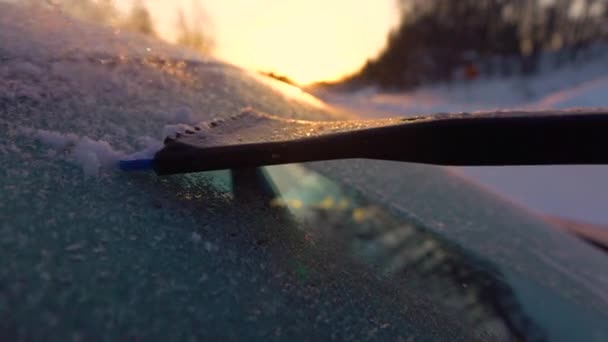 スローモーションクローズアップDof 人は黄金の日の出に車の窓から朝の霜を掃除します 凍った車のフロントガラスから手を削り取り 太陽の上を流れる雪片 霜を取り除くスクレーパー — ストック動画