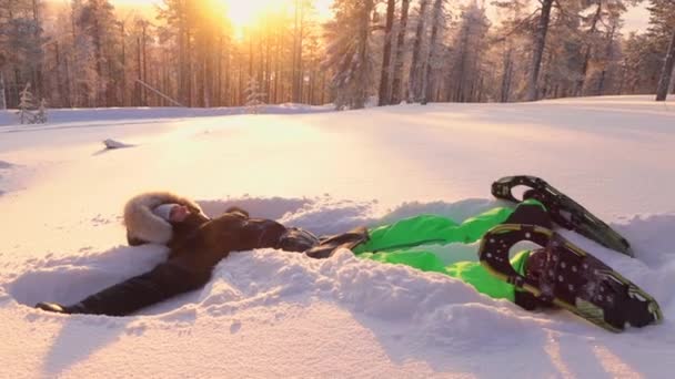 慢动作 欢快的微笑女孩躺在新鲜的粉雪中 在金色的夕阳下 在雪山的山坡上做雪天使 俏皮的女人在阳光明媚的夜晚飞溅雪花 — 图库视频影像