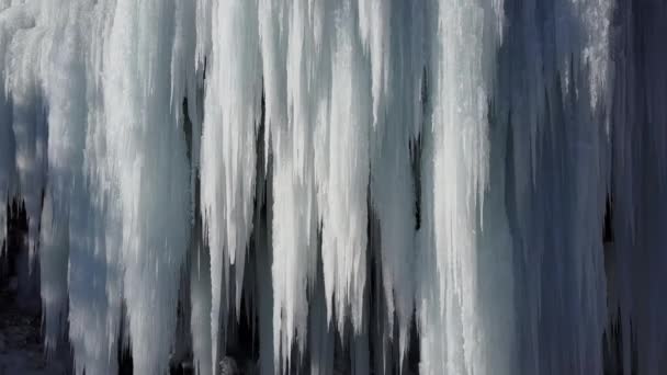 空中クローズアップ冬のカスケードは輝くつららに凍った 冬の日に岩山の崖からぶら下がる見事な凍ったつららら 早春の太陽が凍った滝を溶かす 幻想的な冬の風景 — ストック動画