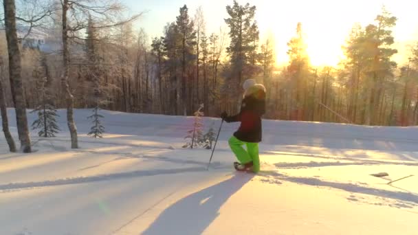 穿着暖冬装的年轻女子在金色的日出时在深雪中穿雪鞋 冬季日落时分 快乐的女孩雪鞋在雪林中徒步旅行 微笑的女性旅行者在寒冬拉普兰锻炼 — 图库视频影像