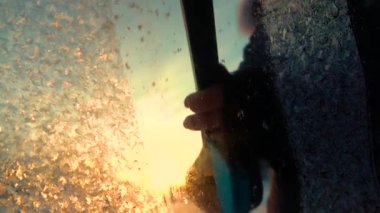 Slow Motion Close Up Dof: Altın gün doğumunda bir araba penceresinden sabah dontemizliği kişi. Donmuş bir arabanın ön camından ses kısıklığı, güneşin üzerinde akan kar taneleri. Kazıyıcı don kaldırma