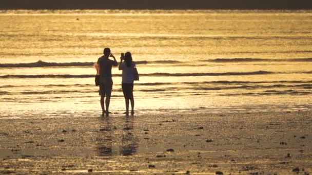 スローモーション シルエット 砂浜で絵のように美しい夕日の写真を撮る認識できない若いカップル ボーイフレンドは日の出時に指でハートの形を作り ガールフレンドはクールな写真を撮ります — ストック動画