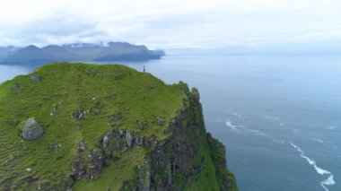 Sonsuz okyanusa bakan kayalık tepede duran tanınmayan kadın fotoğrafçının üzerinden uçuyor. Faroe Adaları 'nda otlak bir tepenin üstünde yürüyüş yapan genç bir kadının muhteşem görüntüsü..