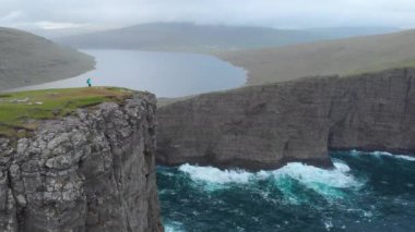 Havadan, Kopya Uzayı: Çimenli bir uçurumun kenarına doğru yürüyüp muhteşem dalgalı deniz ve resimli İskandinav manzarasına bakan tanınmayan genç bir kadın. Faroe Adalarında Turist Yürüyüşü