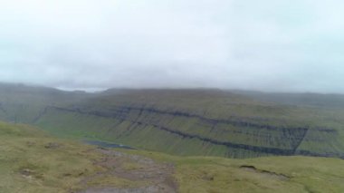 Faroe Adaları 'nda bulutlu bir günde muhteşem bir fiyorda doğru uçmak. Güzel yeşil bir körfez ve göl manzarası nefes kesici. Yoğun bulut tabakası yeşil bir ülkede çarpıcı bir gölün üzerinde toplanıyor.