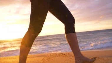 Slow Motion, Lens Flare, Low Angle: Genç bir kadın güneş doğarken çıplak ayakla pikoresk sahilde koşarken havada kumları tekmeliyor. Güzel bir yaz akşamında koşan atletik kız.