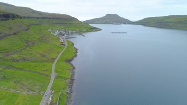 Manzaralı bir yoldan giden araba Faroe Adaları 'nın engebeli yeşil kırsalındaki küçük bir sahil köyüne yaklaşıyor. Çimenli dağların altındaki İskandinav kasabasının muhteşem manzarası.