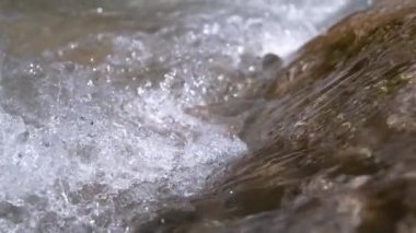 Slow Motion, Macro, Dof: berrak nehir suyu güneşli bir yaz gününde kaygan kahverengi taşların üzerinden akar. Kayalık nehir yatağı boyunca akan parlak soğuk su akıntısının sinematik yakın görüntüsü.
