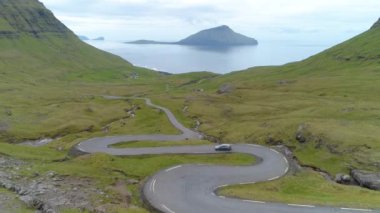 Araba Faroe Adaları kıyılarına ve derin mavi okyanusa doğru dolambaçlı bir kır yolundan gidiyor. Resimli İskandinav manzarasında bir grup turistin arkasında uçuyorlar.