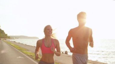Slow Motion, Close Up, Lens Flare: Mutlu genç sarışın kadın ve erkek arkadaşı sakin okyanusun yanındaki manzaralı yolda koşuyorlar. Sıcak yaz güneşi, deniz kenarında birlikte antrenman yapan çiftlerin üzerine parlıyor.
