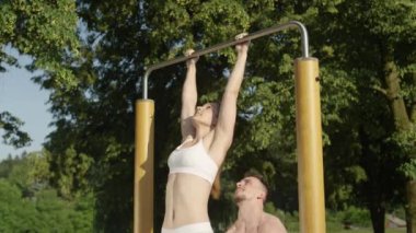 Slow Motion, Close Up: Gülümseyen tişörtsüz koç parkta barfiks çekerken mutlu kız arkadaşını kaldırıyor. Genç beyaz bir çift güneşli doğada birlikte çalışıyor. Egzersiz yapan mutlu adam ve kadın.