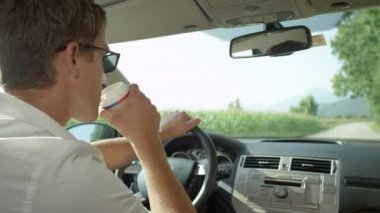 Slow Motion, Close Up, Copy Space: Genç beyaz adam güneşli bir günde boş bir kır yolunda araba sürerken kahve içer. Yolculuktaki yakışıklı adam kağıt bardaktan bir yudum sıcak çay alır.