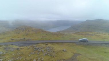 Sisli bir günde, Faroe Adaları 'nın engebeli yeşil kırsalında asfalt bir yolda giden bir arabadan uçarak uzaklaşmak. Tepeleri ve okyanusu aşan yolculukta serin bir araba görüntüsü..
