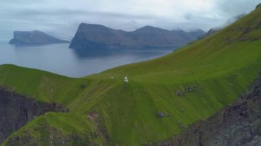 Faroe Adaları 'ndaki çimenli bir tepenin üzerinde beyaz bir feneri ziyaret eden insanların üzerinden uçmak. Yeşil dağlardaki İskandinav adası ve evi etrafında yürüyüş yapan insanların sinematik görüntüleri..