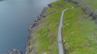 Faroe Adaları 'ndaki boş bir sahil yolunda giden bir arabanın arkasında uçmak. Turistler, İskandinav kırsalındaki siyah kayalık kıyıları ve dik çimenli tepeleri geçerek turneye çıkıyorlar..