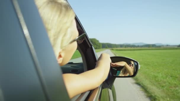 慢动作 特写镜头 一个无忧无虑的白人女孩在风景秀丽的乡间驱车过程中 挥动着她的胳膊 在一个阳光灿烂的日子里 微笑的年轻女子享受着她的暑假之行 — 图库视频影像