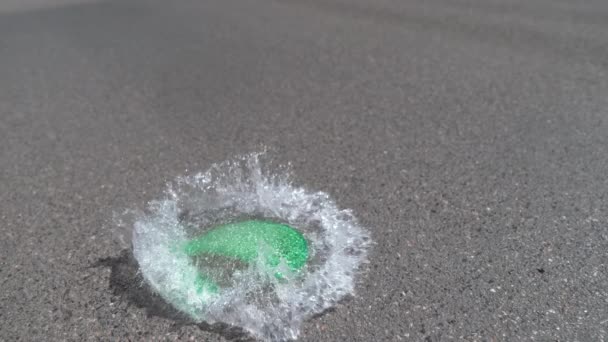 スローモーション クローズアップ 水で満たされた緑の気球はアスファルトの道路に落ち どこでも透明な水を噴霧してポップします 地面への衝撃で爆発する水爆弾のクールな詳細ショット — ストック動画