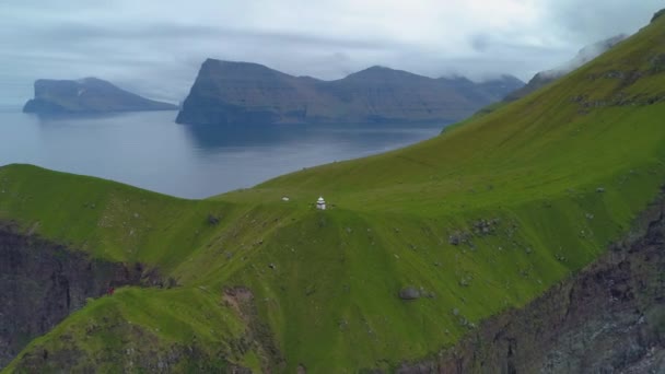 牧歌的なフェロー諸島の芝生の丘の上に白い灯台を訪れる人々の上を飛ぶ 北欧の島や緑の山の中の家をハイキングする人々の映画撮影 — ストック動画