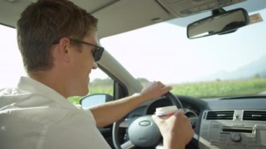 Slow Motion, Close Up: Neşeli genç adam kırsalda araba sürerken kullan-at bardaktan bir yudum sıcak kahve alır. Gülümseyen beyaz erkek, güneşli kırlarda dolaşırken çay içer..