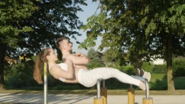 Slow Motion: Mutlu atletik çift birlikte çalışıyor ve açık hava spor salonundaki paralel barlarda mekik çekiyorlar. Neşeli arkadaşlar güneşli parkta antrenman yapıyor. Kız arkadaş ve erkek arkadaş egzersiz yapıyor..