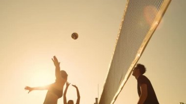 Slow Motion, Low Angle, Close Up, Fens Flare: Gün batımında plaj voleybolu oynarken eğlenen formda gençler. Genç Kafkasyalı kız topu ağların üzerine fırlattı ve adam topu engelledi. Serin yaz sporu.
