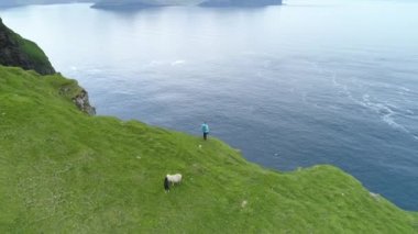 Hava Zaman Bükülmesi: Resimli yeşil Faroe Adaları 'ndaki yüksek bir uçurumdan sakin okyanusa bakan tanınmayan bir kadının üzerinden uçmak. Uzak İskandinav adasındaki engebeli kıyı şeridinin çarpıcı manzarası