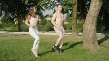 Slow Motion: Genç atletik çift güneşli doğada ısınıyor ve koşuyor. Neşeli beyaz erkek arkadaş ve kız arkadaş parkta egzersiz yaparken eğleniyorlar. Tişörtsüz adam ve formda kadın eğitimi..