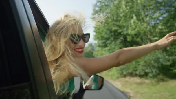スローモーション クローズアップ ケアフリー白人の女の子は 日当たりの良い田舎を巡航しながら 頭と腕を車の窓から出します 陽気ブロンド女性持っています楽しいです中に彼女の夏のロードトリップ — ストック動画