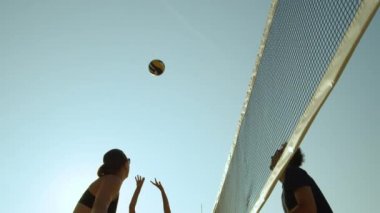 Slow Motion, Close Up, Low Angle: Beyaz adam güneşli bir yaz gününde plaj voleybolu oynayan formda bir kadının vurduğu topu engelliyor. Dişi sporcu topu ağın üzerine fırlatıyor ve uzun adam topu engelliyor..