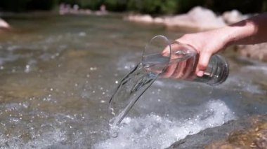 Slow Motion, Close Up: Tanınmayan kadın güneşli yaz gününde berrak nehire cam gibi su döker. Temiz su büyük şeffaf camlardan akarak dağ deresine akar.