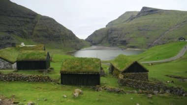 Uçsuz bucaksız vadide çimlerle kaplı geleneksel İskandinav evleri. Faroe Adaları 'ndaki küçük bir köyün her iki tarafında yükselen dik dağların nefes kesici görüntüsü.